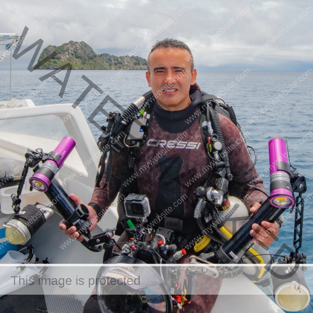 Photograph of Edwar Herreno, sitting in a boat withhis dive gear in front of the ocean backdrop that features a little island. Promoting artist gallery MIRAME Fine Art.