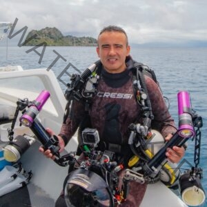 Photograph of Edwar Herreno, sitting in a boat withhis dive gear in front of the ocean backdrop that features a little island. Promoting wildlife photographer on MIRAME Fine Art.