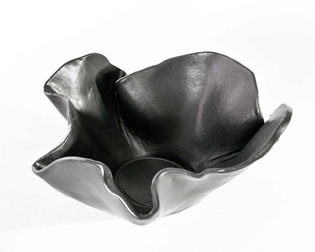 From the "Black Magnolia" series, a lustrous black ceramic bowl with undulating, petal-like edges gleams against a white backdrop. , an original work available at MÍRAME Fine Art.