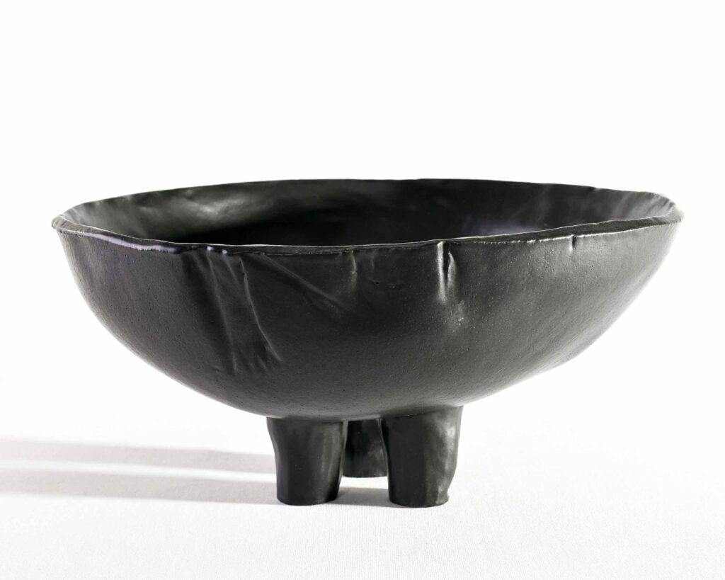 This "Black Magnolia" bowl, with a matte, textured finish and organic rim, rests on three legs against a softly shadowed backdrop. , an original work available at MÍRAME Fine Art.
