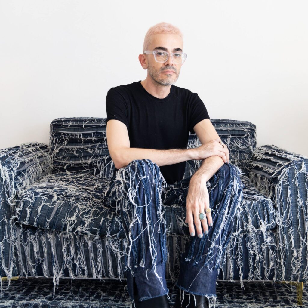 An individual with bleached hair and glasses, composed on a frayed denim chair, exudes quiet poise against a stark white backdrop. , an original work available at MÍRAME Fine Art.