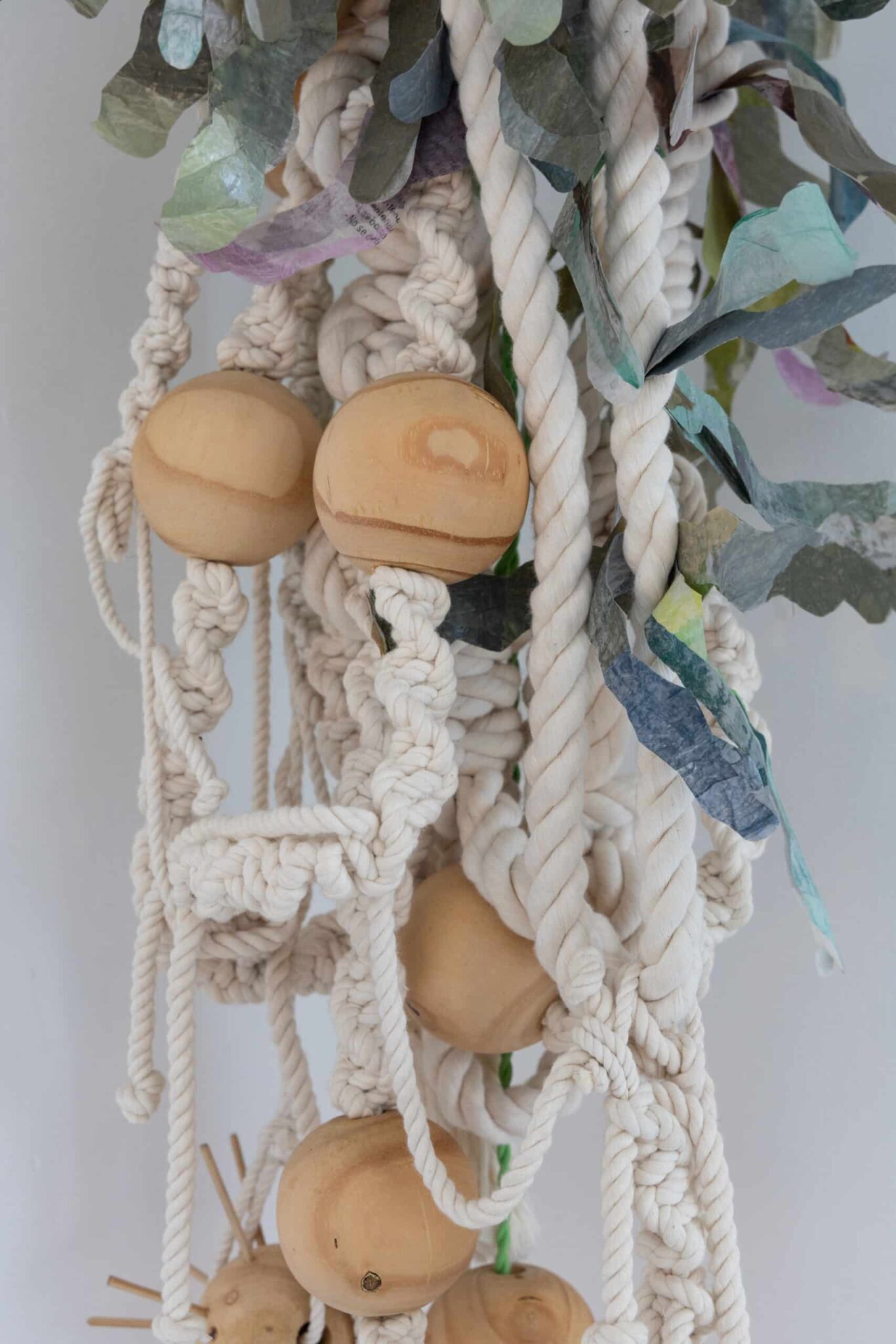 This detailed view of the Olimpia macramé reveals intertwined white rope, wood beads, and pastel leaves beside an Art Deco vase. , an original work available at MÍRAME Fine Art.