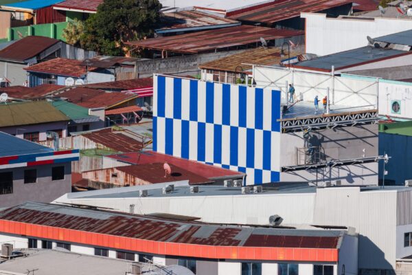 Urban image by photographer Leonardo Ureña, focusing on the rooftops of San José. A blue and white striped strip stands atop the rooftops. Part of his rooftop photography series. Part of the San José Art Guide.