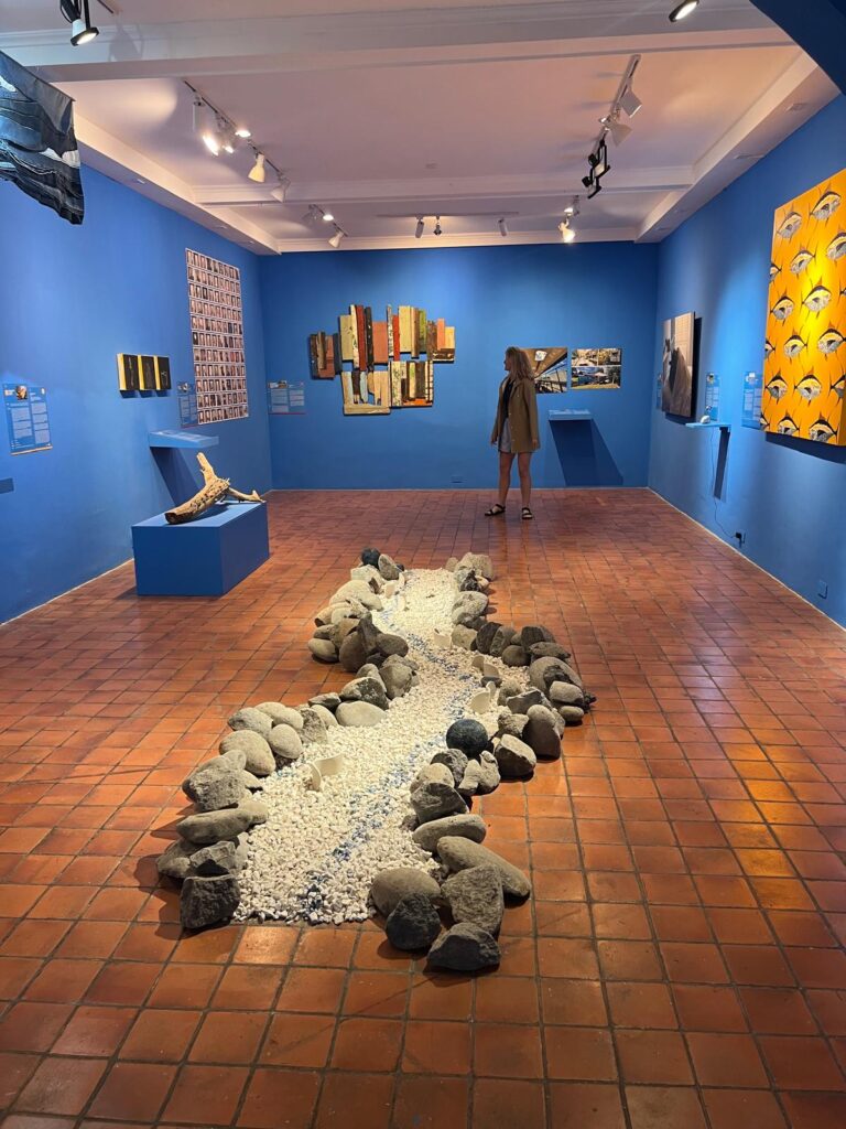 Art exhibition with blues and different types of art hanging. A central sculpture is on the floor and a woman is walking through the space.