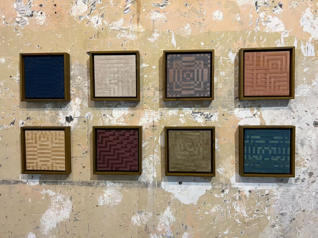 Eight small colourful textile artworks hanging on a wall, featured in Más Allá Del Índigo at San José's MADC. Costa Rica's Contemporary Art Scene.