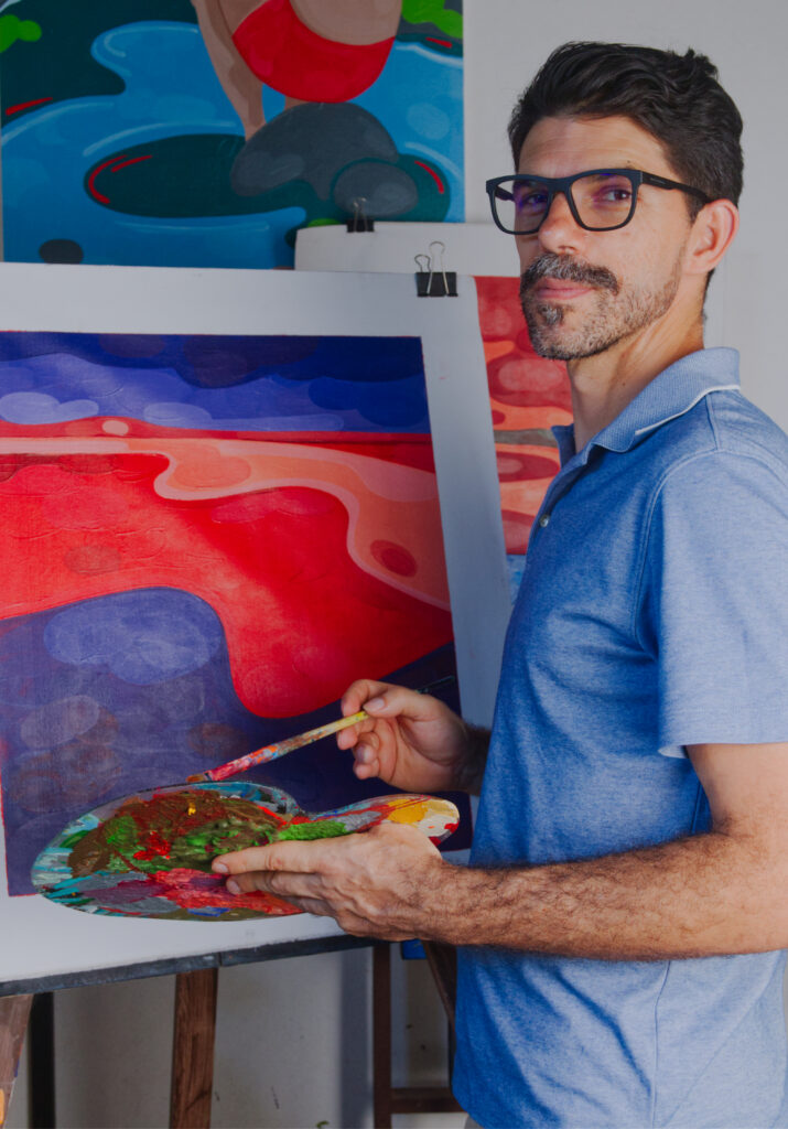 Artist standing beside his paintings with a paintbrush in his hand.