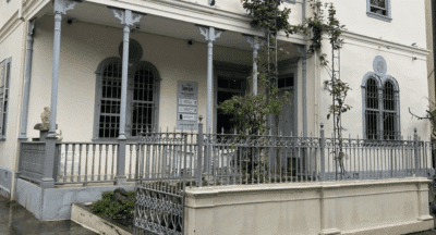Outside of an art gallery building, white paint and thin vertical columns. Featuring an exhibition of local Costa Rican artists.