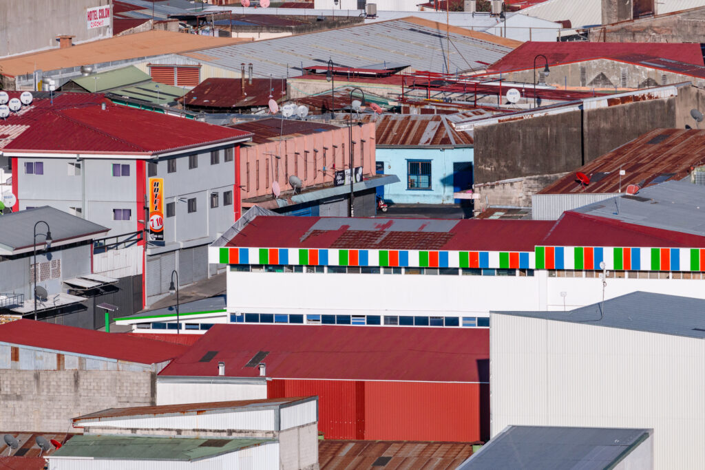 Part of Leonardo Ureña's Urban Series. A tightly framed urban scene in San José, Costa Rica, showing a dense patchwork of rooftops in shades of red, rust, and grey. In the mid-ground, a white building stands out with a brightly striped red, green, and blue horizontal trim. Details such as hotel signs, satellite dishes, streetlamps, and mismatched windows hint at the area’s mix of residential and commercial use. A narrow strip of concrete street with a motorbike is partially visible, blending into the surrounding architecture.