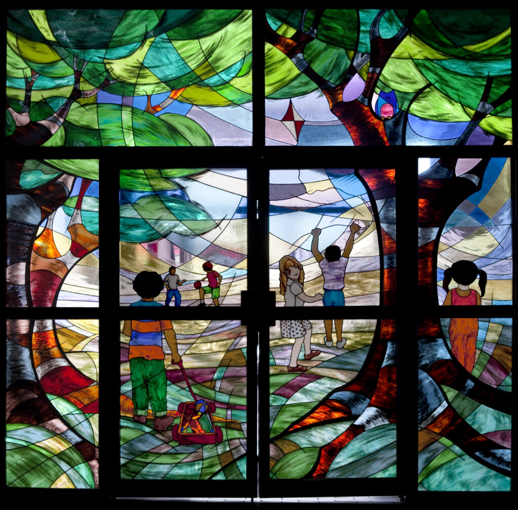 Colourful stained glass children scene, with children running around playing underneath green trees.