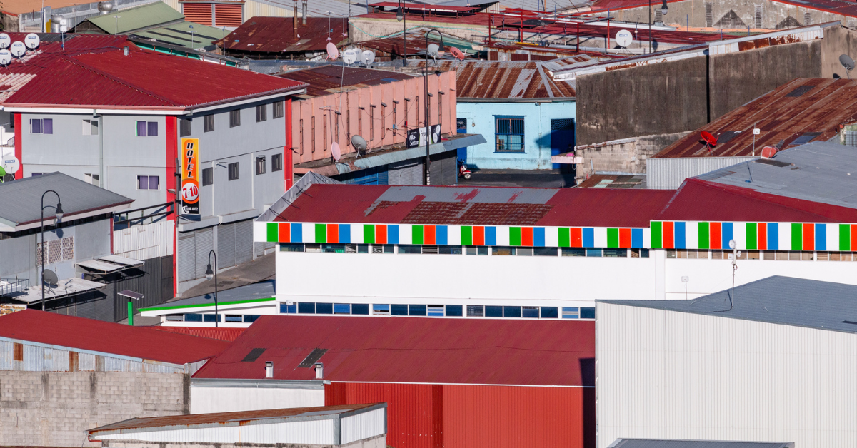 Part of Leonardo Ureña's Urban Series. A tightly framed urban scene in San José, Costa Rica, showing a dense patchwork of rooftops in shades of red, rust, and grey. In the mid-ground, a white building stands out with a brightly striped red, green, and blue horizontal trim. Details such as hotel signs, satellite dishes, streetlamps, and mismatched windows hint at the area’s mix of residential and commercial use. A narrow strip of concrete street with a motorbike is partially visible, blending into the surrounding architecture.