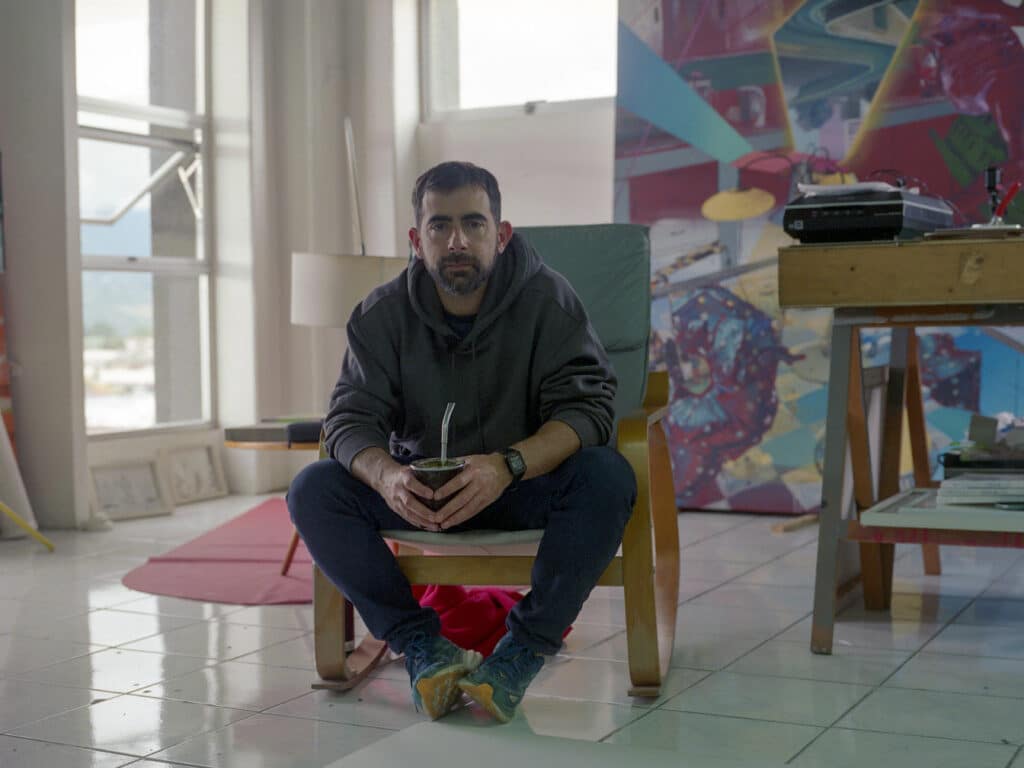 Costa Rican artist Luciano Goizueta seated in his studio, holding a mate gourd. He sits on a wooden chair surrounded by canvases, a desk with art materials, and large abstract paintings leaning against the walls.