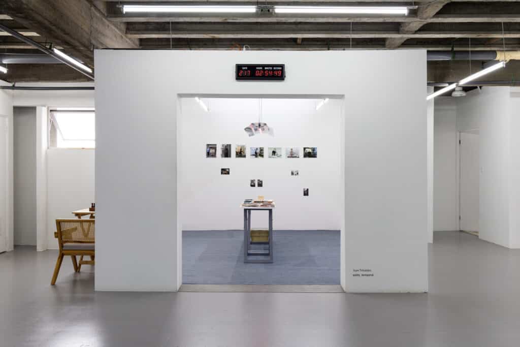 Installation view at Salita.temporal in San José, where Costa Rican photographer Juan Tribaldos began his residency and launched his photobook project Solve et coagula. A minimalist gallery room features small photographs arranged on the wall, a central display table with prints and books, and a countdown clock above the entrance.
