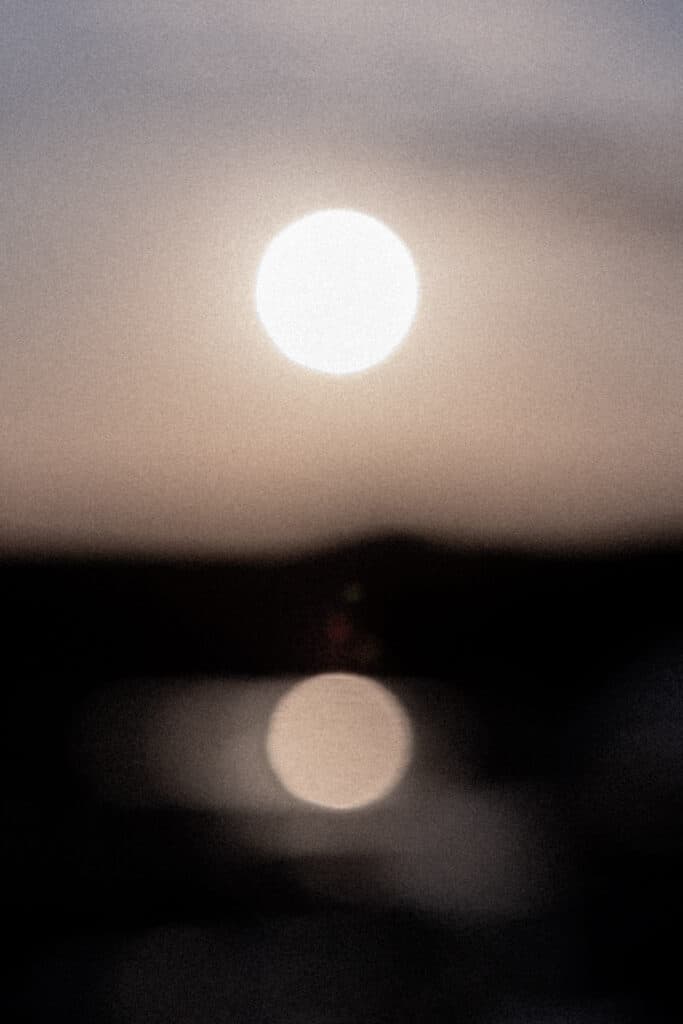 A softly blurred photograph of the sun above the horizon, with its reflection visible on dark water below. The image, in muted tones of beige, grey, and black, creates an abstract, atmospheric effect typical of Juan Tribaldos’s Costa Rican photographer contemporary photographic style.