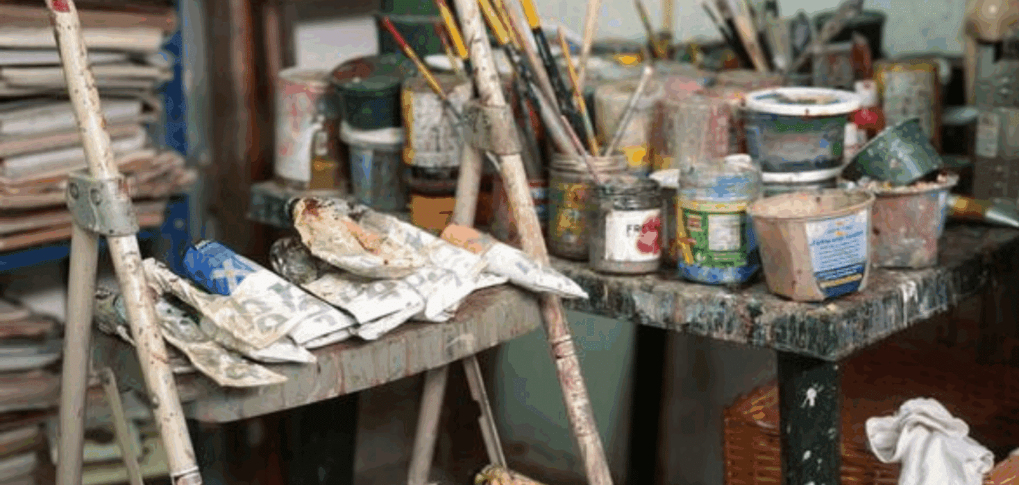 A studio scene photographed by a Costa Rican photographer, showing paintbrushes in jars, squeezed paint tubes, and a splattered step stool surrounded by art supplies, capturing the working environment of an artist.