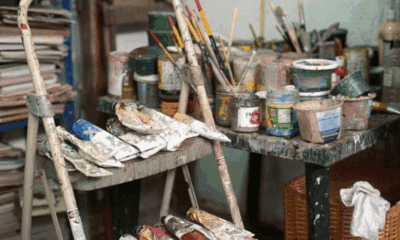 A studio scene photographed by a Costa Rican photographer, showing paintbrushes in jars, squeezed paint tubes, and a splattered step stool surrounded by art supplies, capturing the working environment of an artist.