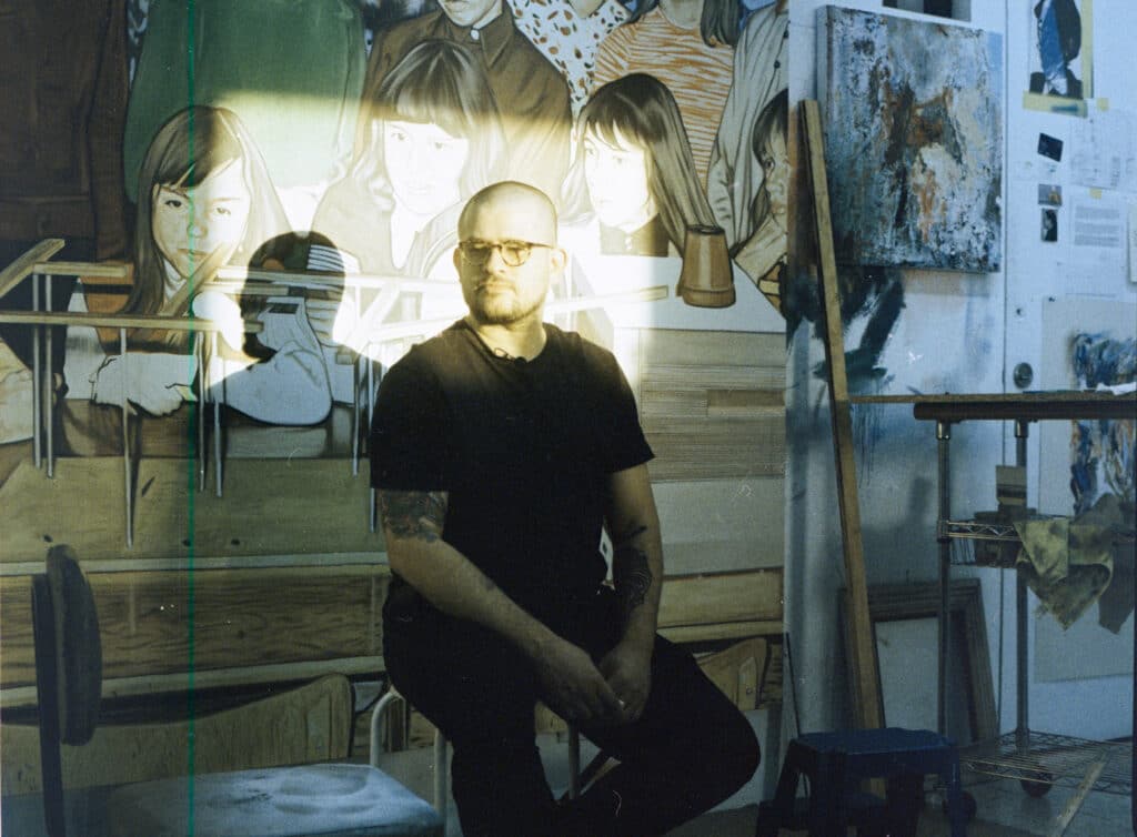 Costa Rican artist Emmanuel Rodríguez Chaves seated in his studio, illuminated by a shaft of light. Behind him, large figurative paintings of children and works in progress cover the walls, alongside sketches and notes.