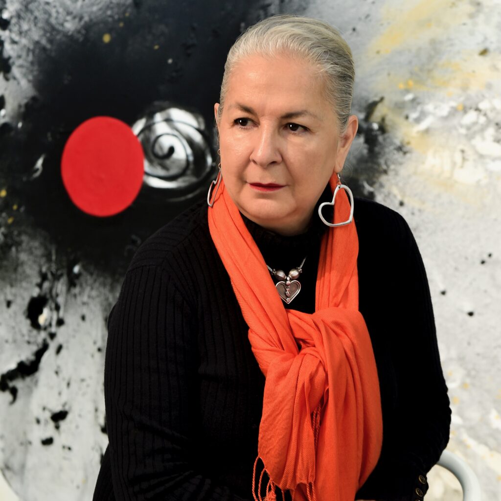 Portrait of the artist Rossella Matamoros in her studio, dressed in black with bright orange scarf in front of one of her abstract paintings.