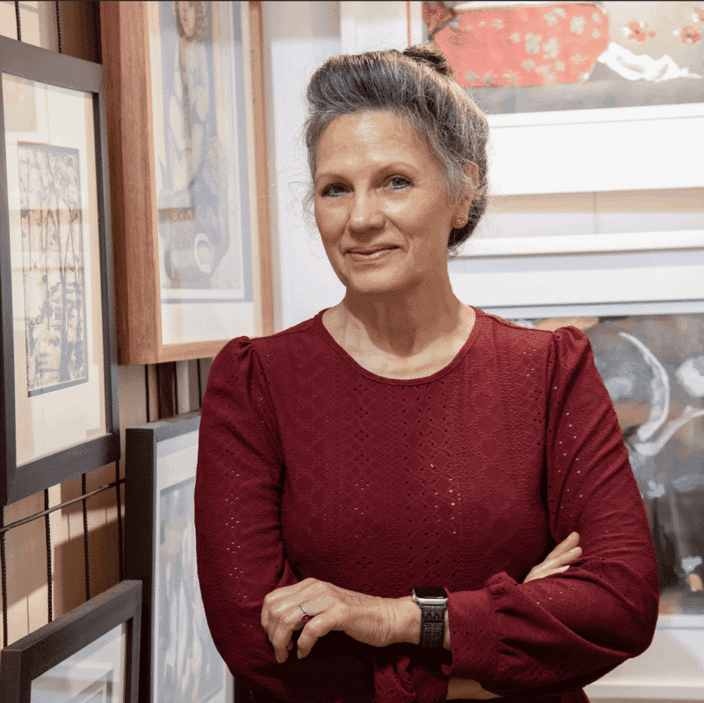 portrait of artist Lorena Villalobos in her studio, standing in front of her paintings. She is wearing a deep red long-sleeved top and stands with crossed arms.