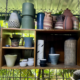 Ceramic vessels by Gema Ossenbach displayed on studio shelves in Tres Ríos, Costa Rica, including stoneware vases, pitchers and bowls in terracotta, sage green and deep grey glazes, with tropical foliage visible behind.