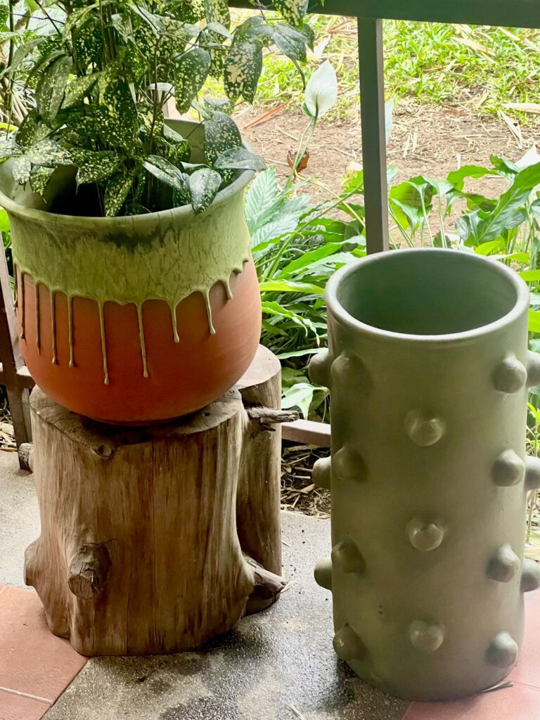 Two ceramic pieces by Gema Ossenbach: a terracotta planter with pale green drip glaze displayed on a wooden stump, alongside a matte sage green cylindrical vase with raised spherical detail, photographed in her Tres Ríos studio garden.