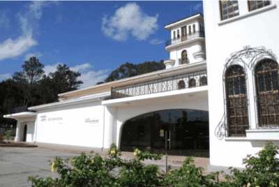The Museo de Arte Costarricense, a white modernist edifice with arched windows and an elegant tower, stands amid lush gardens beneath a radiant blue sky. Showcasing selected Costa Rican art exhibitions in the county.