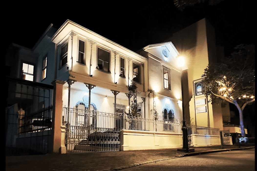 An elegant, two-story Costa Rican landmark, bathed in warm and blue light, foregrounded by art signage and softly lit foliage. It is Galería Talentum, the location of exhibition Patria e Identidad.
