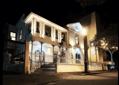 An elegant, two-story Costa Rican landmark, bathed in warm and blue light, foregrounded by art signage and softly lit foliage. It is Galería Talentum, the location of exhibition Patria e Identidad.