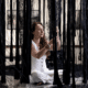 Seated cross-legged, a woman - Alessandra Sequeira - in a white jumpsuit intricately weaves black cords amid loose threads in a refined mixed media installation. Promoting her exhibition at the Juan Santamaría Historical and Cultural Museum. , an original work available at MÍRAME Fine Art.